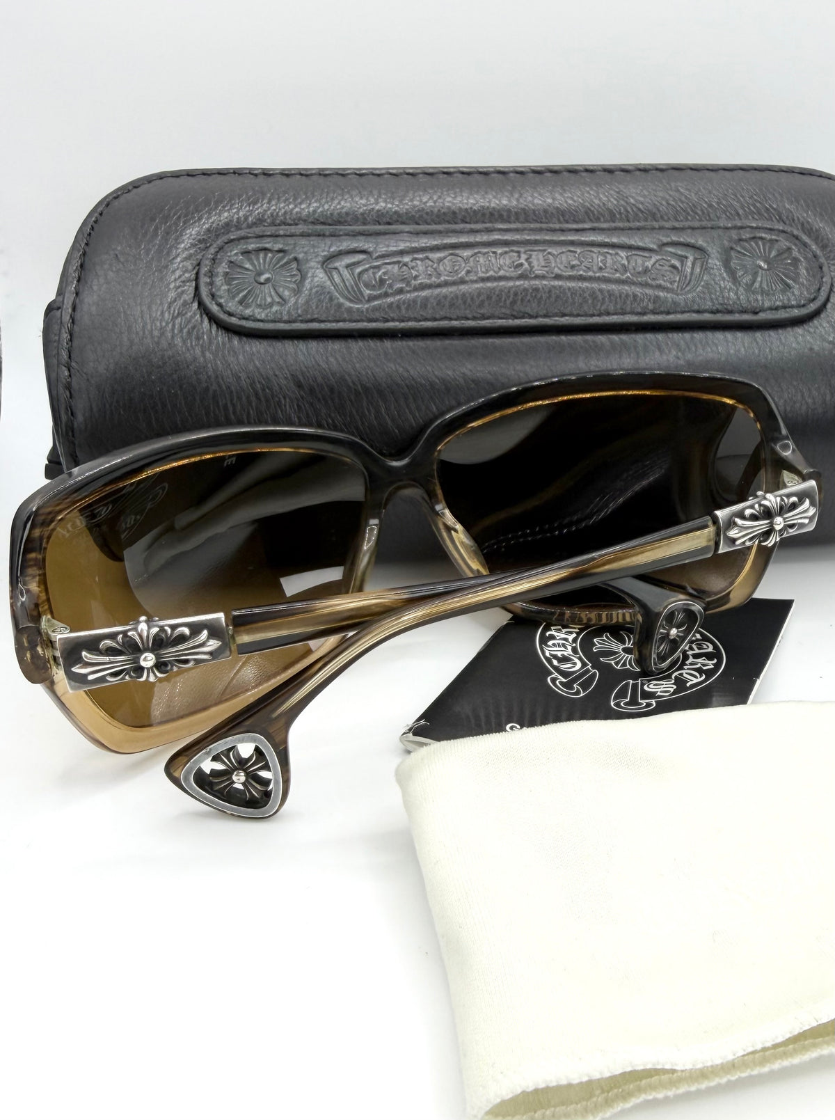 A pair of vintage-style Chrome Hearts sunglasses with tortoiseshell frames and dark lenses, displayed with a black leather case, a white chrome hearts cleaning  cloth.
