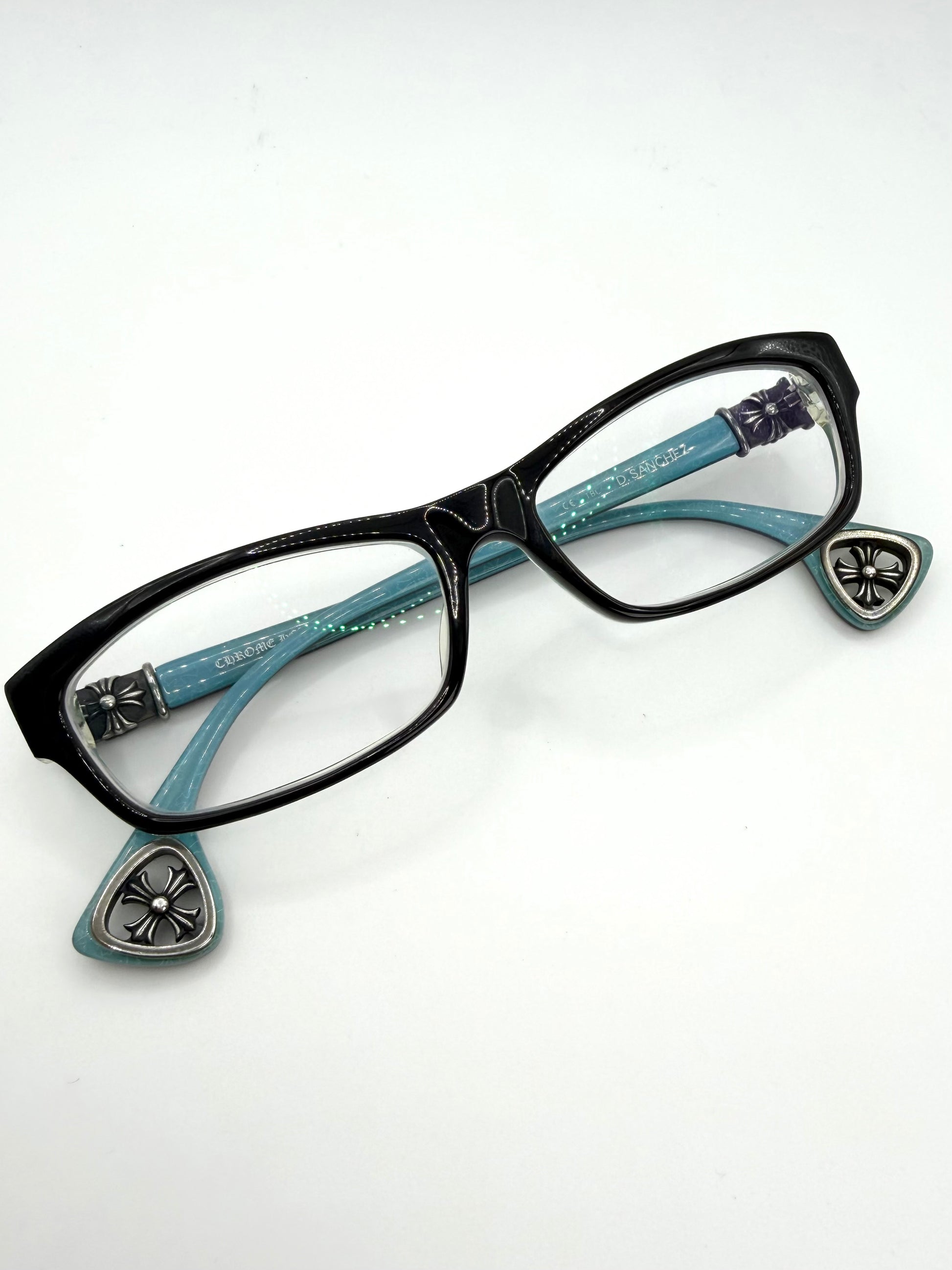 Black chrome hearts d Sanchez cat-eye eyeglasses with turquoise blue temples featuring silver-tone decorative metal crosses and cut-out cross temple tips, shown on a plain white background.