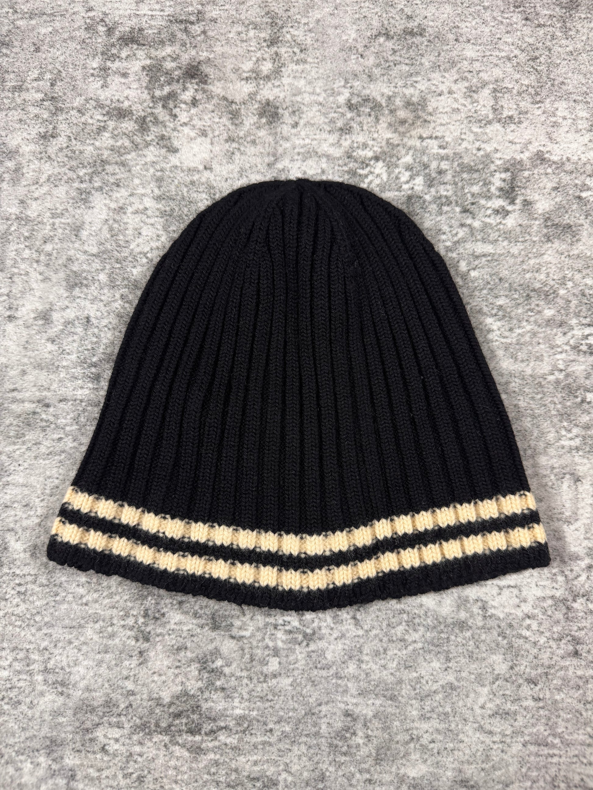 Junya Watanabe Comme des Garçons Man ribbed knit beanie in black with cream contrast stripe, photographed flat on a textured grey surface.