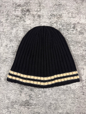 Junya Watanabe Comme des Garçons Man ribbed knit beanie in black with cream contrast stripe, photographed flat on a textured grey surface.