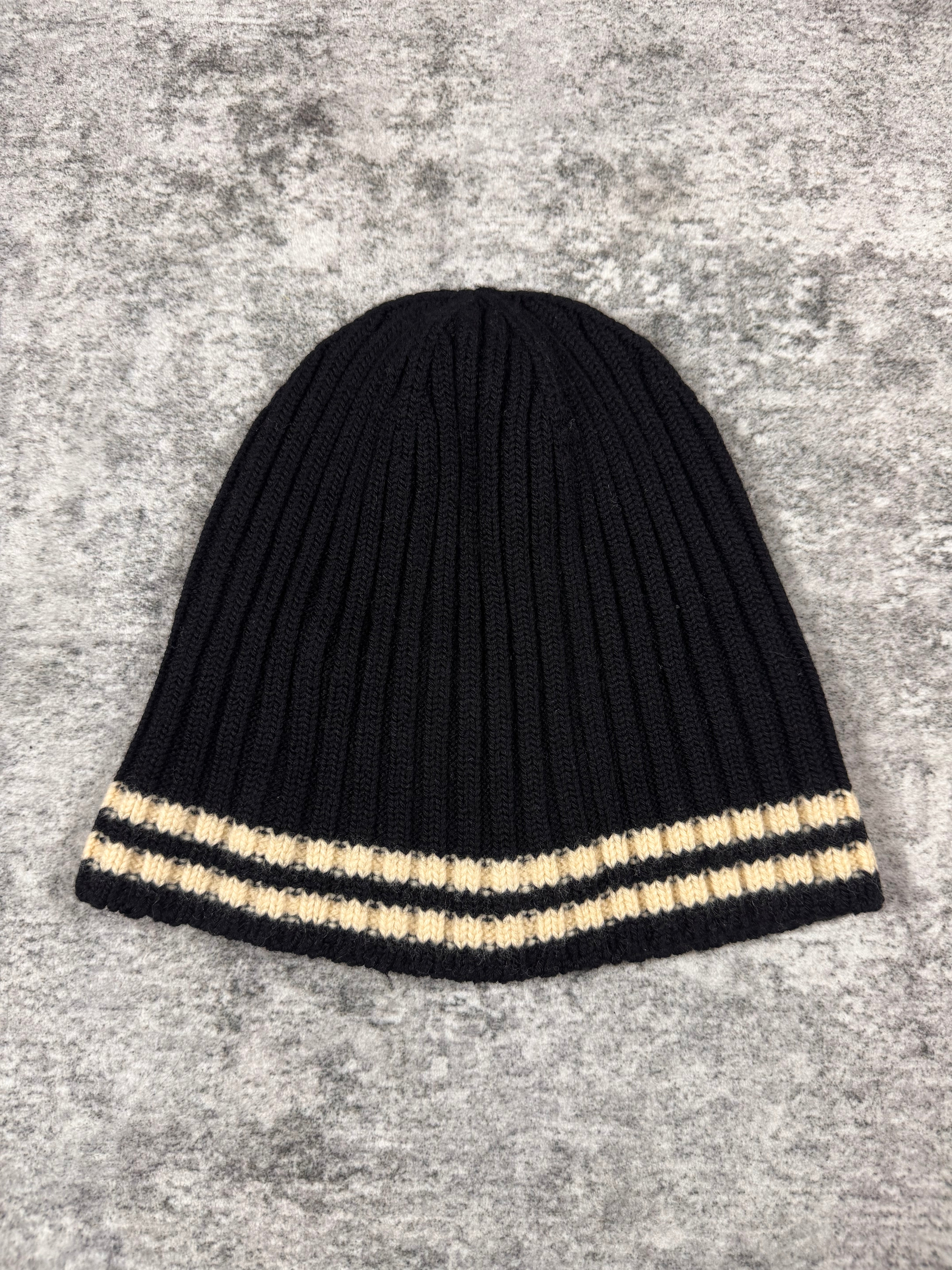 Junya Watanabe Comme des Garçons Man ribbed knit beanie in black with cream contrast stripe, photographed flat on a textured grey surface.