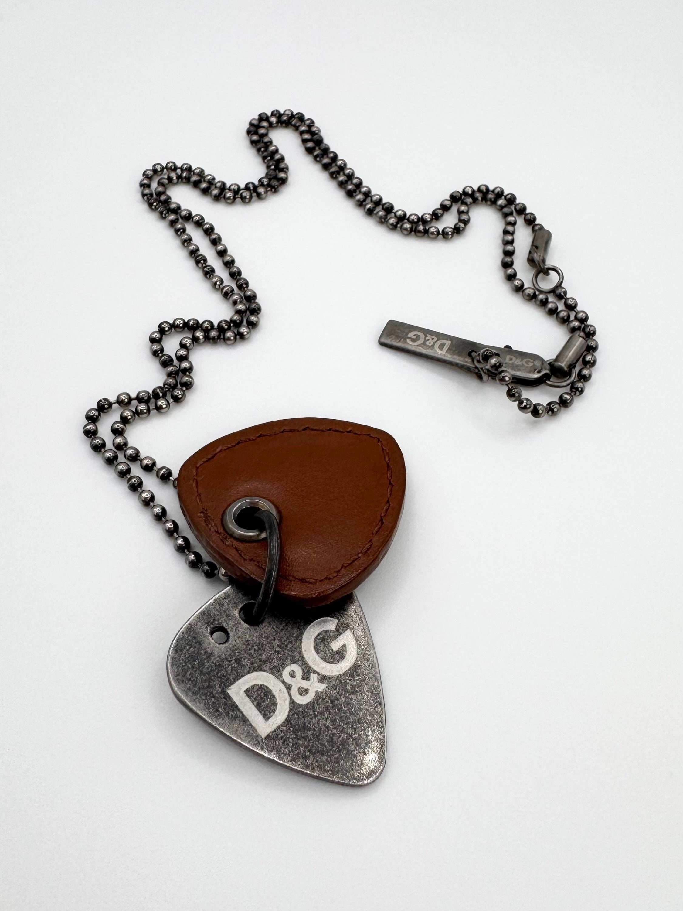 Vintage Dolce & Gabbana D&G guitar pick pendant necklace with brown leather tab and dark silver-tone ball chain on white background.
