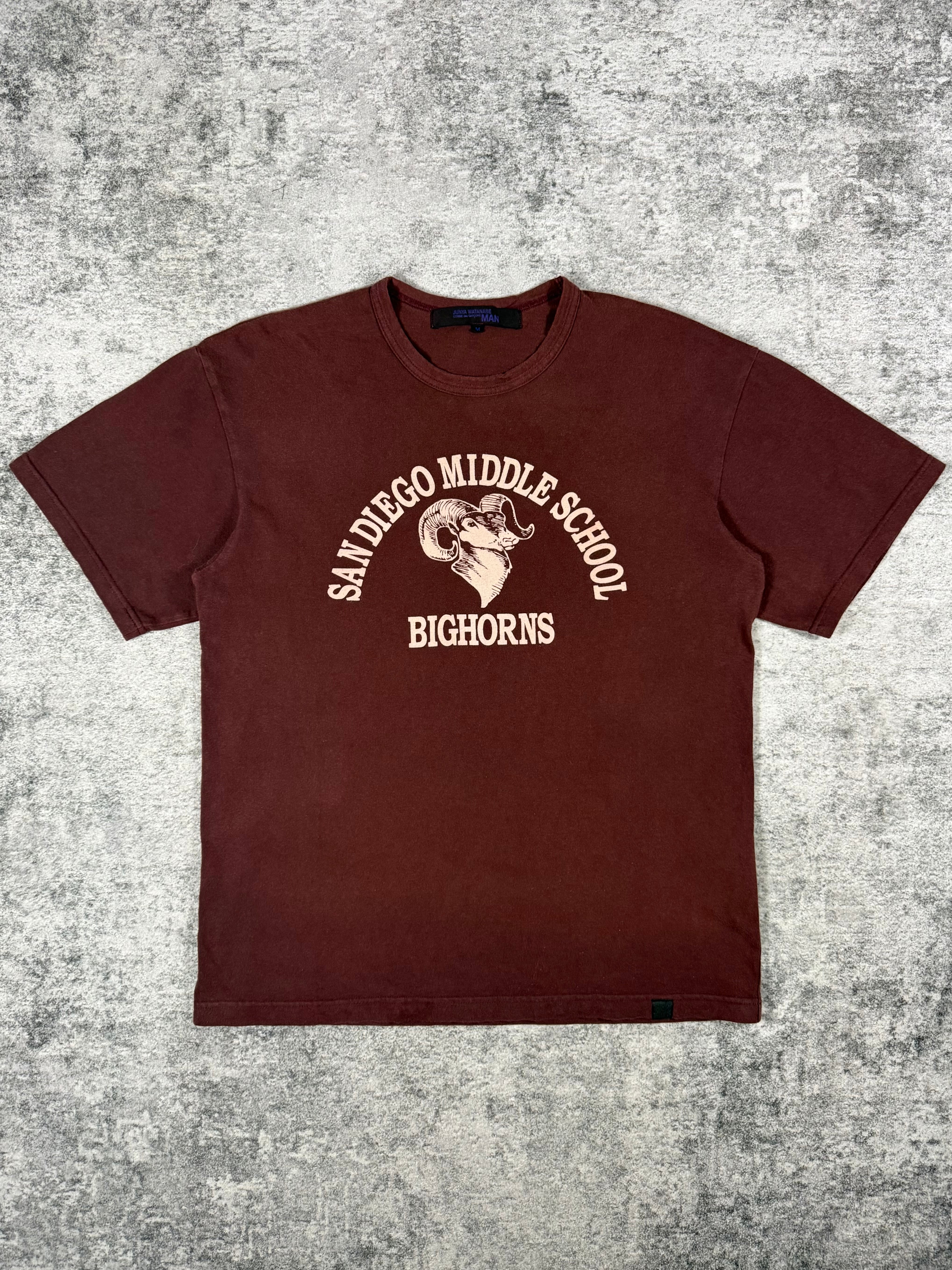 Archival Junya Watanabe American collegiate school wear inspired t shirt featured the San Diego Middle School Big Horns graphic with a ram centered on a burgundy cotton t shirt showcased on a grey gradient carpet. 