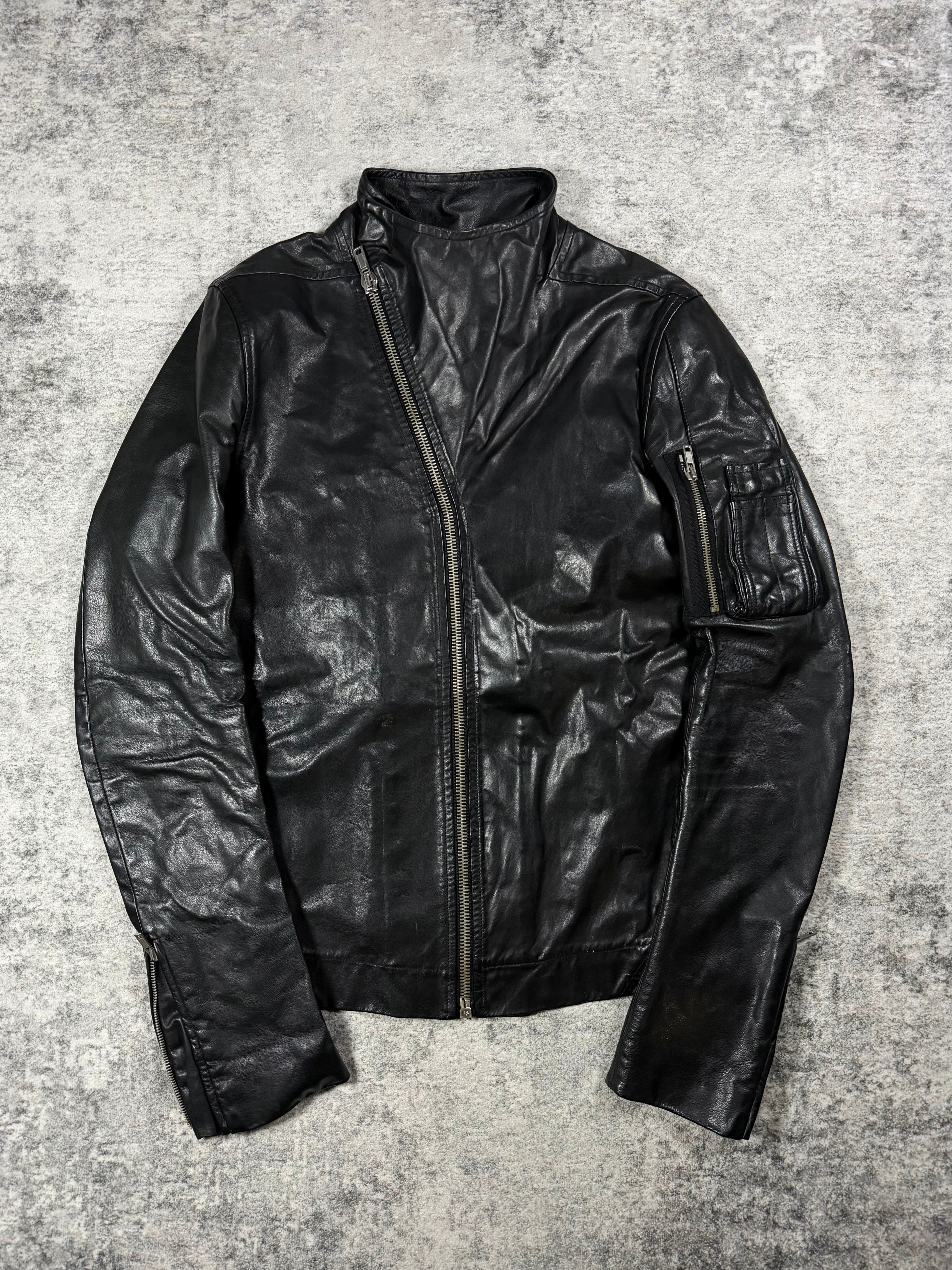 A black Rick Owens leather jacket leather jacket with a high collar and asymmetrical zipper, laid flat on a textured gray concrete background.