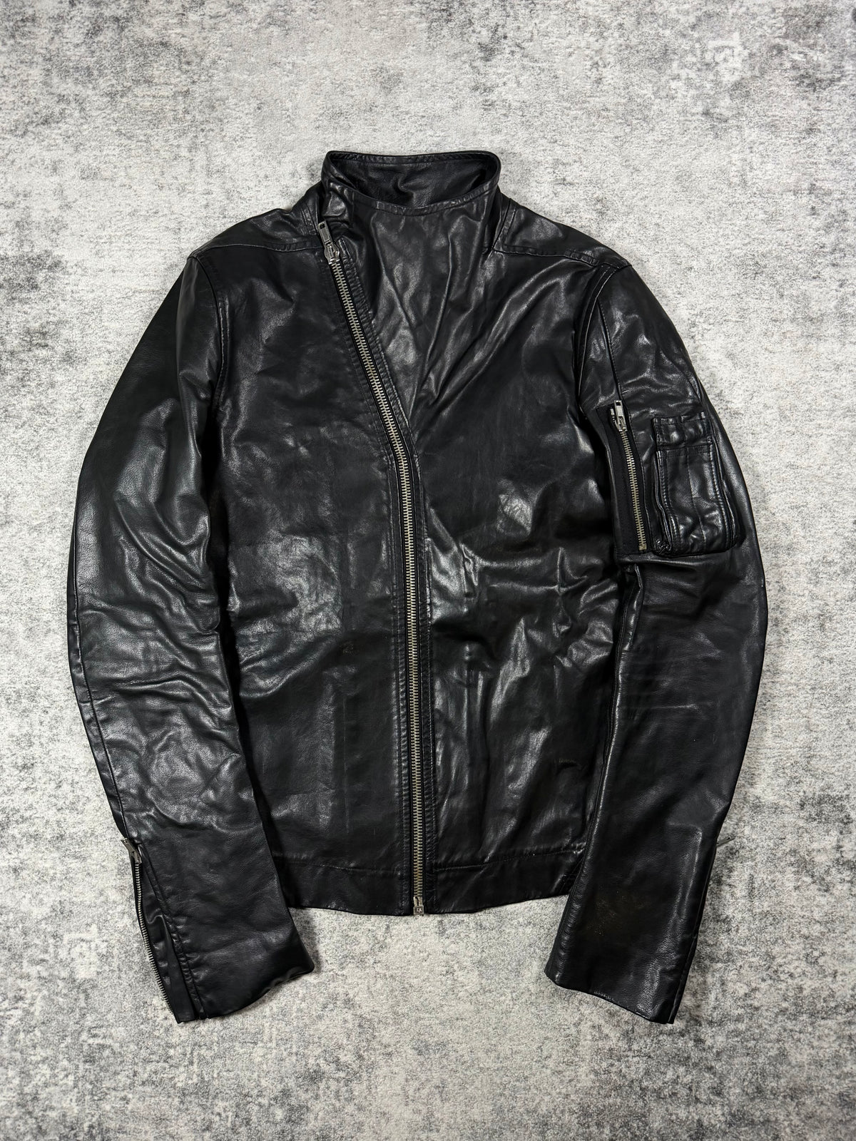 A black Rick Owens leather jacket leather jacket with a high collar and asymmetrical zipper, laid flat on a textured gray concrete background.