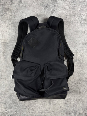 A black Undercover canvas backpack with multiple front utility pockets and padded shoulder straps, laid flat on a textured gray concrete surface.