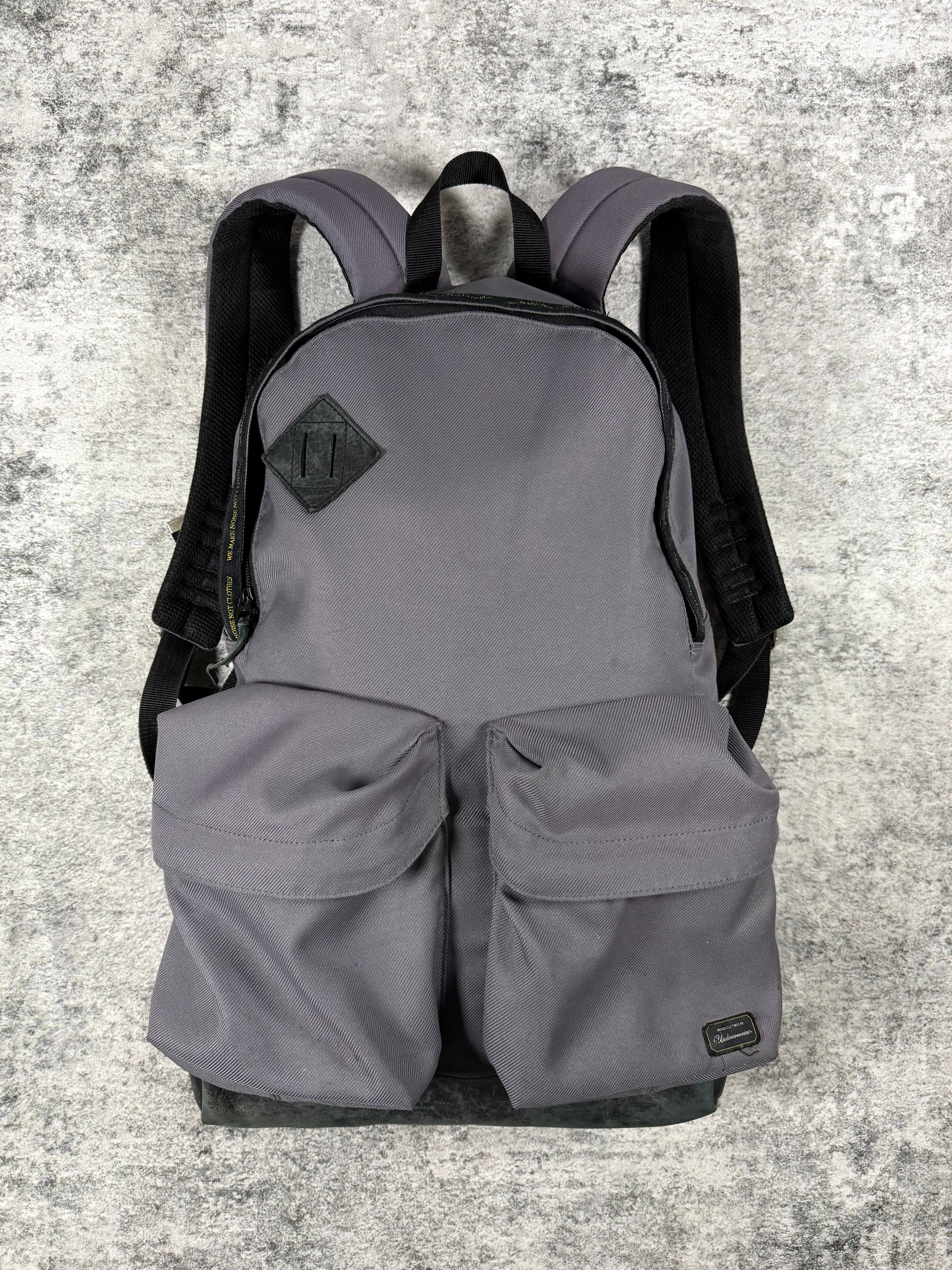 A gray Undercover backpack from 2009 with black shoulder straps and a black diamond-shaped logo patch on the front, displayed against a textured gray concrete background.