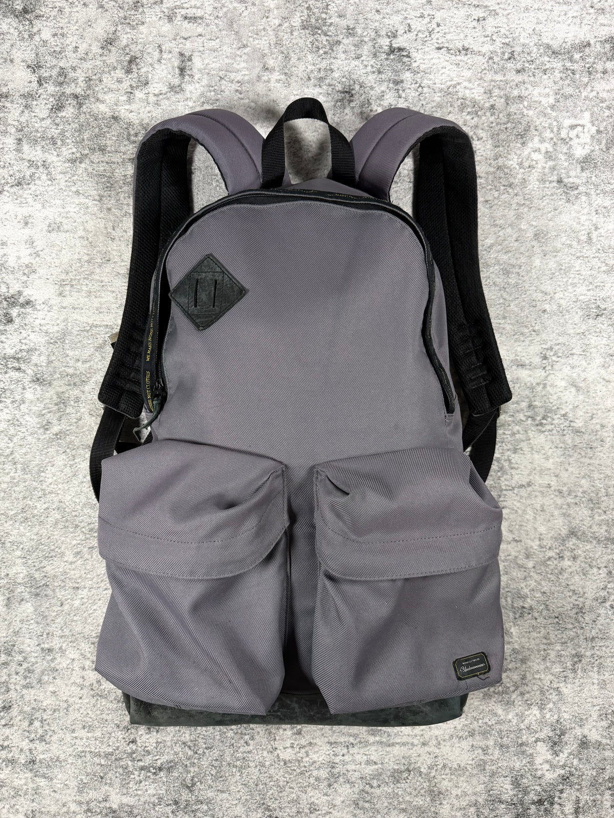 A gray Undercover backpack from 2009 with black shoulder straps and a black diamond-shaped logo patch on the front, displayed against a textured gray concrete background.