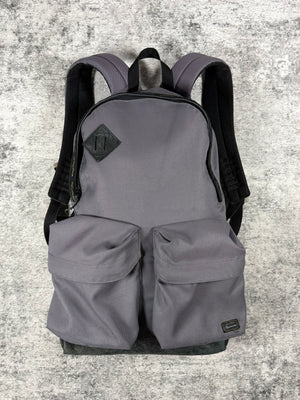 A gray Undercover backpack from 2009 with black shoulder straps and a black diamond-shaped logo patch on the front, displayed against a textured gray concrete background.