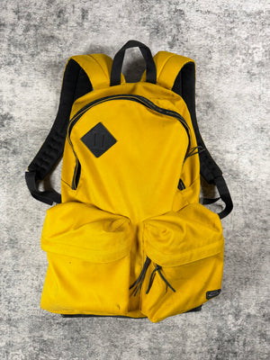 A bright yellow Undercover backpack from 2009 with black straps, zippers, and a black diamond-shaped logo patch on the front, laid flat on a textured gray carpet surface.